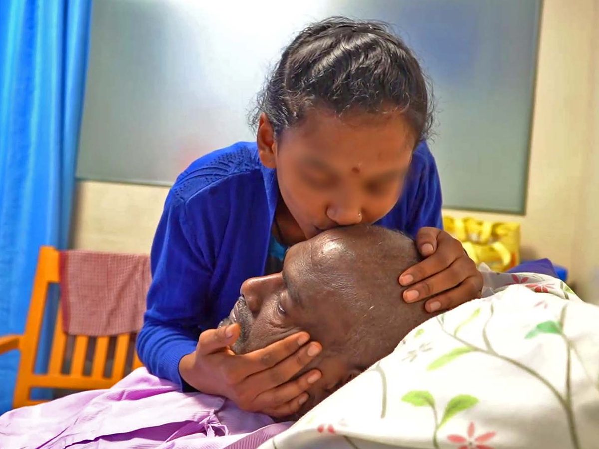 A young woman in a blue cardigan leans over a hospital bed to kiss the forehead of a man resting in it
