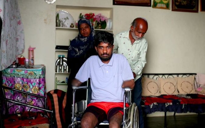A young man seated in a wheelchair at home, flanked by an older woman in a hijab and an older man standing close behind him