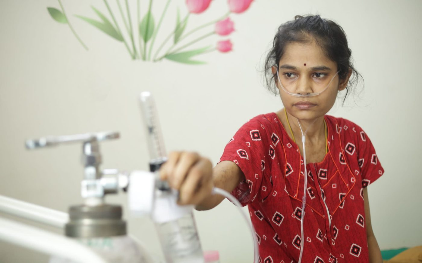 A young woman wearing a nasal cannula in a hospital room, her hand on the flowmeter of an oxygen cylinder